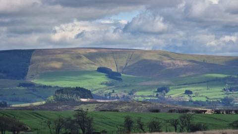 White and grey clouds with some light blue sky sit above fields in the foreground and mountains on the horizon