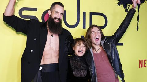 Finalists Sam Wells, Mai Whelan, and Phill Cain attend Netflix's "Squid Game: The Challenge" finale watch party at Terminal 5 on December 06, 2023 in New York City. Sam is a tall white man with short brown hair and a long brown beard. He wears an unbuttoned black blazer exposing a bare chest and midriff. He holds his right hand up in a celebratory fist. Mai stands next to him and is much shorter, barely as tall as Sam's shoulder. She has a short brown bob and wears a sparkly black top. On her left is Phill, about a head taller than Mai. He wears a black leather jacket over a maroon T-shirt and has shoulder length brown hair. He also holds his left arm aloft and all three are smiling for the cameras. They're pictured at a screening event with a yellow background emblazoned with Netflix's logo