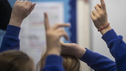 Young children with their hands up in a classroom. They they wearing dark blue jumpers and white shirts