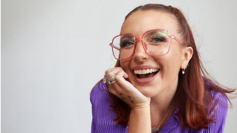 Ellie Middleton smiling at the camera, with her head resting on her hand. She is wearing pink glasses and a purple shirt.