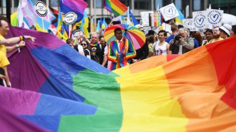 People walk behind Pride rainbow flag carrying placards and flags