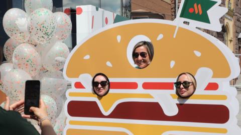 A woman takes a photo of three women posing in a board model of a giant hamburger at a Vkusno I Tochka restaurant in Moscow