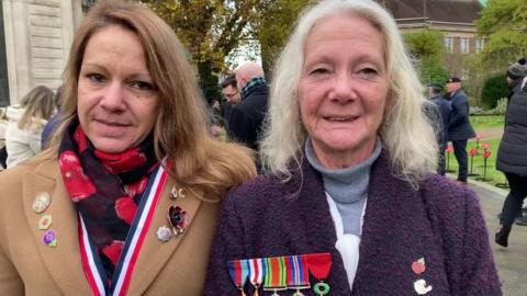 Woman wearing brown coat with one medal stands next to older woman wearing blue coat and several medals