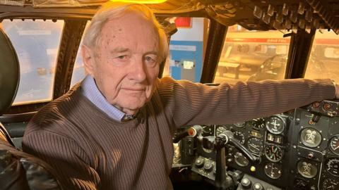 A man in a brown jumper sitting in a cockpit of a plane