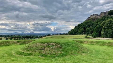 Cloudy grey skies with some small areas with sun breaking through over a grassed field with a huge mound in the foreground