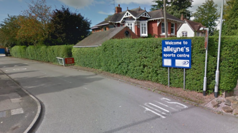 The entrance to Alleyne's sports centre includes a road going from the bottom right to the left of the photo and a blue sign with white letters saying "Welcome to alleyne's sports centre".