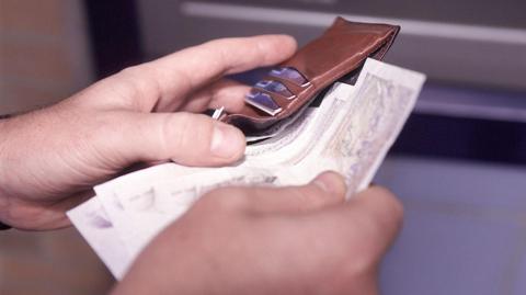 A man's left hand holds a leather wallet while the right hand slips bank notes out of it