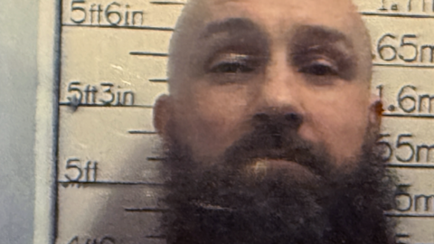 Michael Ross. he has a bald head and a big brown, bushy beard. He is standing in front of a height chart and looking at the camera.