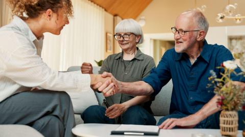 Financial advisor, handshake and old couple with documents for insurance, saving or planning. Retirement, shaking hands and person with senior man, woman and paperwork for mortgage or pension in home - stock photo