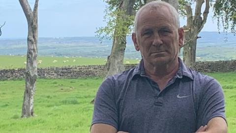 Ian Wilkinson is looking at the camera with his arms crossed. He is wearing a grey t-shirt and has short white hair. He is standing at the front of his home. There is a short stone fence behind him. Sheep are feeding in the grass field behind the fence.