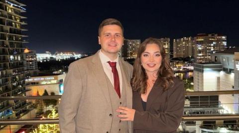 Mathew is wearing a tanned brown suit and Kylie is wearing a dark brown blazer behind a skyline of apartment blocks.