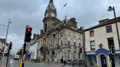 Kendal Town Hall in Kendal's town centre. There are people milling about outside the building, which has a large clock tower.