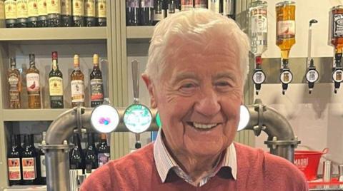A man with white hair and wearing a jumper is smiling at the camera at a bar.