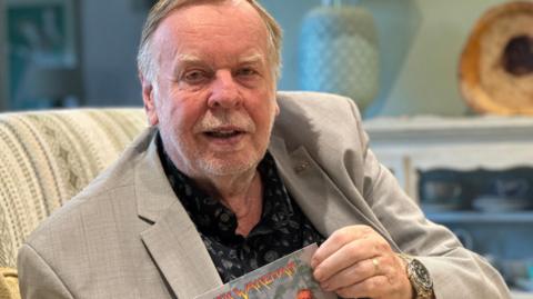 Rick Wakeman, who has short fair hair and beard and is wearing a light coloured suit jacket over a black shirt.