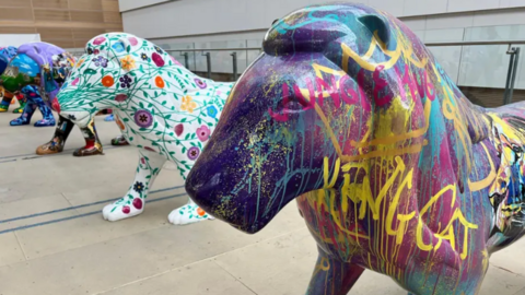 Three painted lion sculptures, one painted in graffiti and another in flowers, on display on a paved area. Other lion sculptures can be seen behind them.