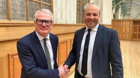 Two men wearing suits shake hands, standing in a wood-panelled room. Both of grey hair and one is wearing thick black glasses.