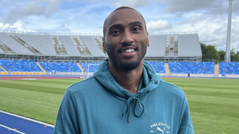 A man wearing light blue is looking at the camera, standing on a blue athletics track. Blues seats and grey seats in a stand are in the background.