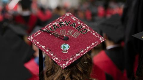 A red graduation cap on the head of a graduate with long brown hair, pictured from the back. On the cap is printed "Harvard 2025" and "Learn to change the world". Other graduates with red gowns can be seen blurred in the background