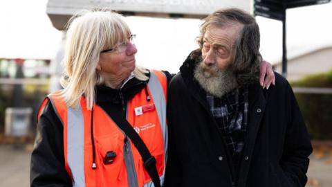 A lady with blonde hair and wearing glasses, with her arm around an older man with grey hair and a beard. They are looking at each other. 