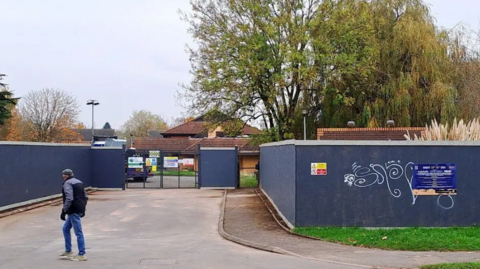 The site of the former Three Counties Hotel. It is boarded off with blue boards as a man walk past the entrance.