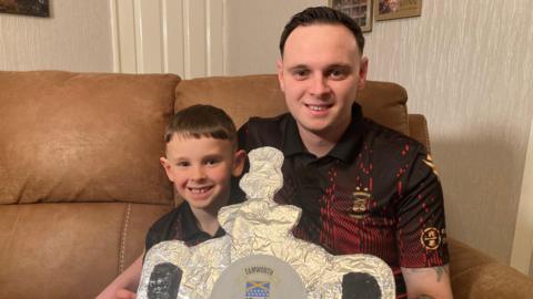 A young boy and a man are sat on a brown sofa, wearing matching football tops and holding a trophy-shaped piece of tinfoil.