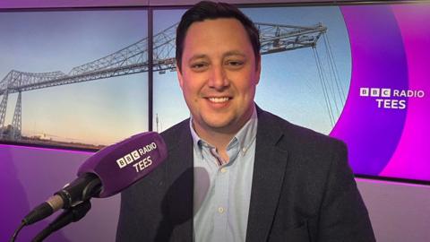 Ben Houchen sitting in the BBC Radio Tees studio with a purple mic in front of him and two screens behind him showing a picture of Middlesbrough's Transporter Bridge and the radio station name and logo. He is smiling at the camera and wearing a light blue shirt and dark suit jacket.