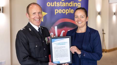 Chief Supt Roy Linden in police uniform, South Devon Police Commander, (left) giving a certificate to Det Ch Insp Becky Davies wearing a navy blue shirt and blazer. Both are smiling. Behind them is a sign which says "outstanding people". The wall behind it is cream. 