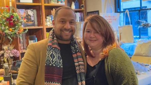 A man and a woman stand smiling inside a shop. Shelves behind the counter are full of books, tote bags and bunches of flowers.