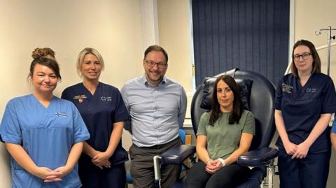 Emma Cullen who has dark hair and is wearing a green top sat on a hospital chair with hospital staff stood beside her