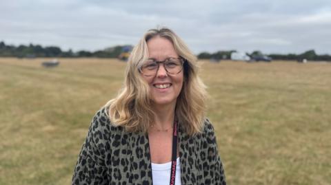 A woman, who has shoulder-length blonde hair, is wearing tortoiseshell glasses and an animal-print cardigan and white T-shirt and has a black and pink lanyard around her neck. She is standing in a field with trees and greenery in the far distance.