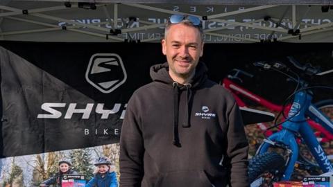 A man with short dark hair has sunglasses on his head and is wearing beige trousers and a black hoodie. He is standing in front of a black gazebo with the word "Shyre" on the top in large lettering. On a backdrop it says "Shyre bikes" and has a photo of a red kids bike and a blue kids bike.