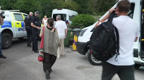 The backs of people walking towards a white van or minibus, with a blue and yellow marked police vehicle parked up.