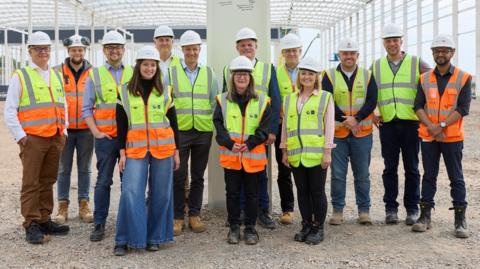 A large group of people standing in the middle of a construction site, all wearing white hard hats, and high-viz jackets, smiling and looking straight at the camera. A white steel post is behind them and scaffolding. 