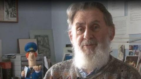 John Cunliffe is pictured in a room surrounded by books and photographs. He has a white beard and there is a Pat figure in the background.