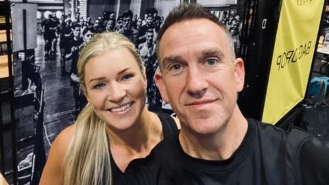 A woman with long blonde hair, and a man with short dark hair both look directly into the camera. There is a large black and white image of a group of people behind them