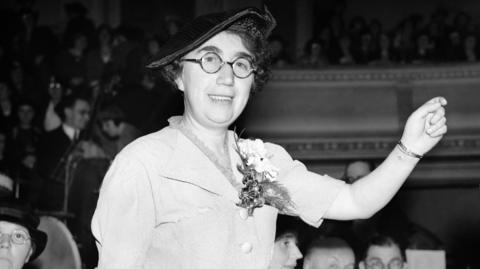 A black and white photo of Florence White. She is wearing a light coloured jacket and a dark hat. She has her left arm raised as if to make a point.