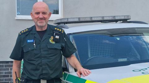 The image shows a paramedic stood in a the usual NHS ambulance service dark green uniform standing next to an emergency vehicle. He is looking straight at the camera and smiling while resting his hand on the car. 