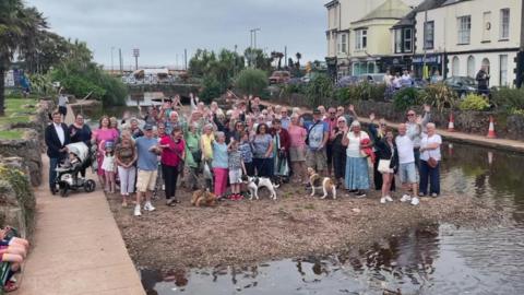 A large group of people standing on a silt bank in Dawlish Water. Some of them have dogs on leads. Some people have their arms in the air