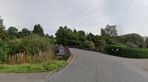 Sebergham Bridge is a stone walled bridge with greenery on either side. A sign to the left of the bridge reads Sebergham.
