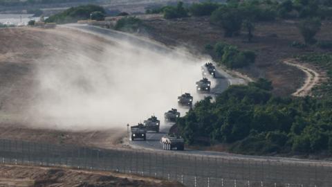 An Israeli military convoy manoeuvres near the Israel-Gaza border. Armoured vehicles can be seen driving along a winding road, with greenery to the right and sand to the left. A fence can be seen in the foreground.