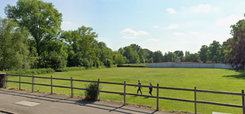 A large green field with a border of trees and bushes. two people walk over the grass. in the foreground is wooden fencing slightly higher than the field. on the other side of it is a concrete path and a road 