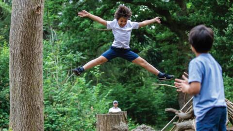 A young boy wearing blue shorts and a white t-shirt doing a splits jump with his arms out, above several large tree stumps in the middle of the forest. There is another young boy to the right clapping his hands together and watching