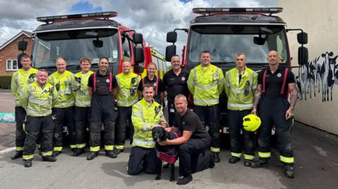 A group photo of firefighters in their uniforms. In the front of the group is a black labrador. They are all standing in front of two fire engines.