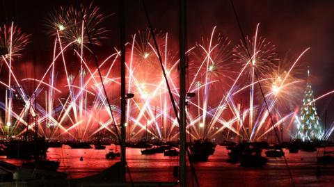 A bright display of pink and yellow fireworks shooting up in the sky in the shape of cross crossing fountains. There are boats and a harbour in the foreground at a decorated Christmas tree glowing with white lights to the right.