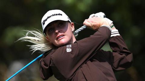 Charley Hull swinging a blue golf club on the course while wearing a white cap, white glove in her left hand and a brown jacket