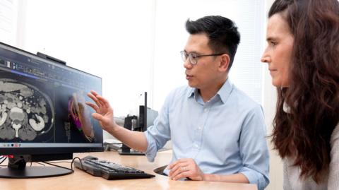 Surgeon Mr Siong-Seng Liau and Jane Gardner looking at a 3D scan on a computer screen in an office. He has dark hair and spectacles and she has long, dark hair.