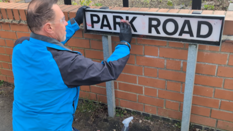 A man wearing a blue coat using a wipe to clean a road sign stating Park Road