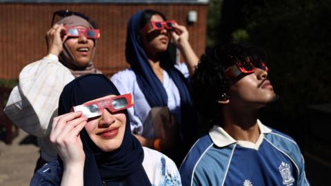 A group of people look up at the sky wearing solar glasses outside Greenwich Observatory