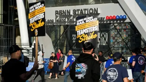 Commuters exit a train station as striking rail workers hold up signs