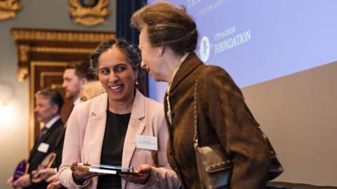 Princess Anne standing on stage next to a woman in a black dress and pink blazer, accepting an award. The princess has short brown hair and wears a brown top with a brown handbag over her left shoulder. 